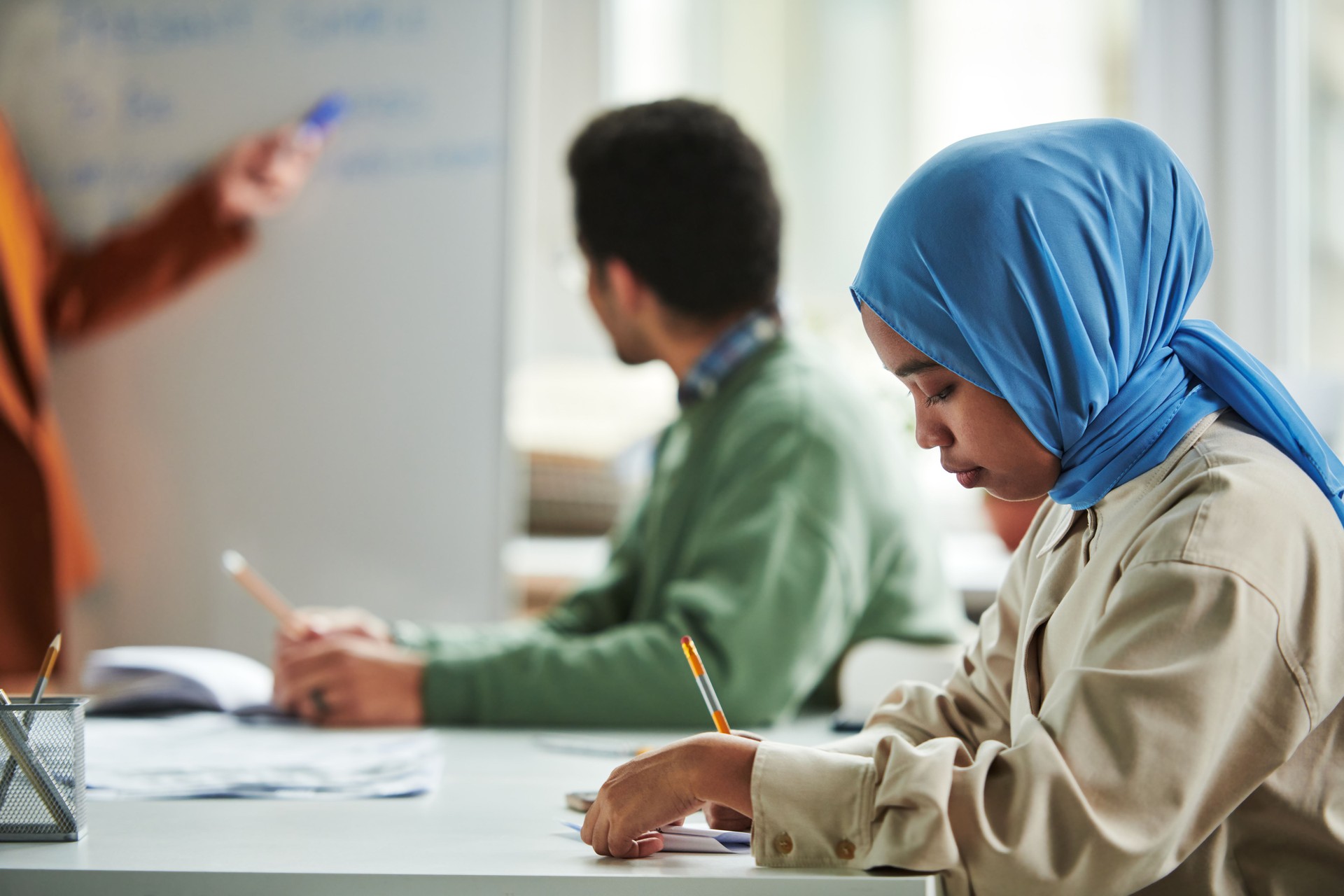 Side view of young serious Muslim woman in blue hijab making notes